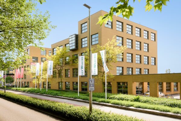 Kantoorgebouw aan de Rivium 2e Straat met vlaggen van Van Waning, omringd door groen en een heldere blauwe lucht.
